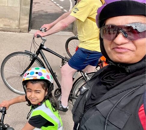 Usma is posing in a selfie with a young child, riding their bicycles and wearing safety helmets.