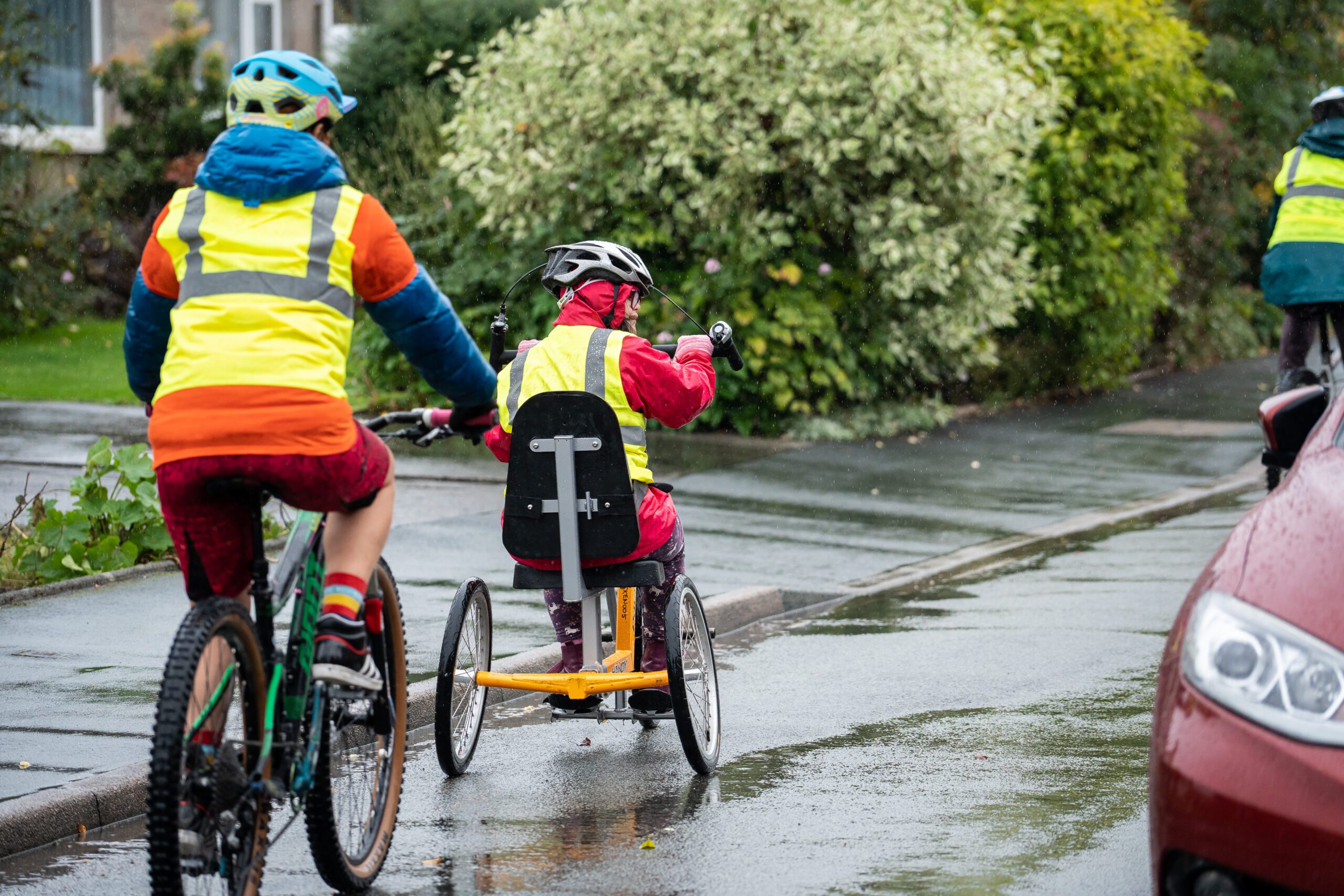 A Bikeability instructor and student cycling on the road. The student is riding a hand tricycle. They are both in hi vis and wearing helmets.