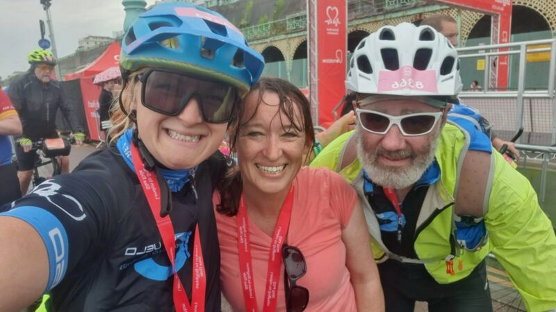 A selfie of Emma, Emily and Adrian at the finish line of the London to Brighton charity cycle ride. They are all grinning.