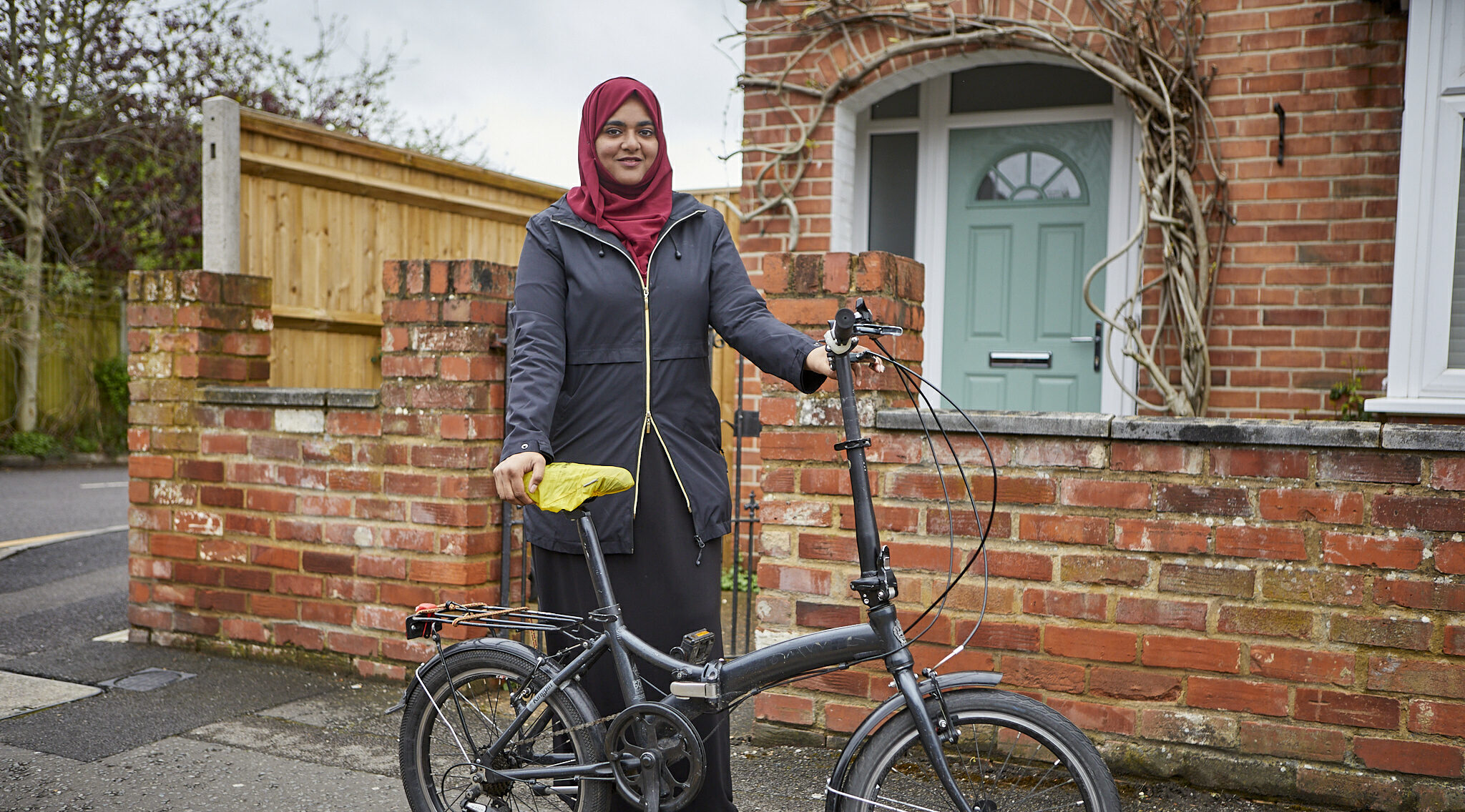 A woman in a red head scarf in standing in front of a house with a folding cycle
