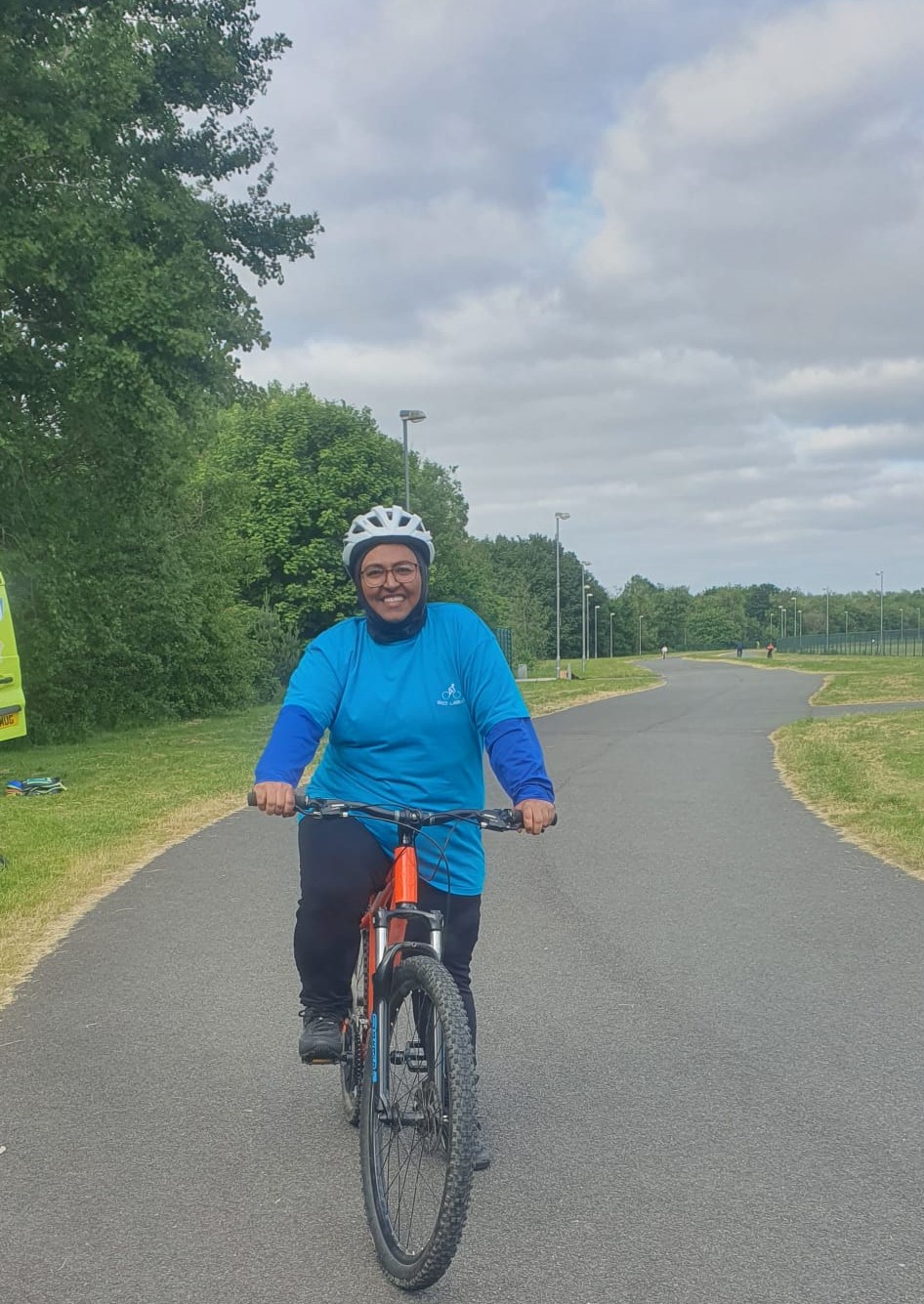 Naida smiling on her bicycle. She is wearing a bright blue t shirt and a white helmet.