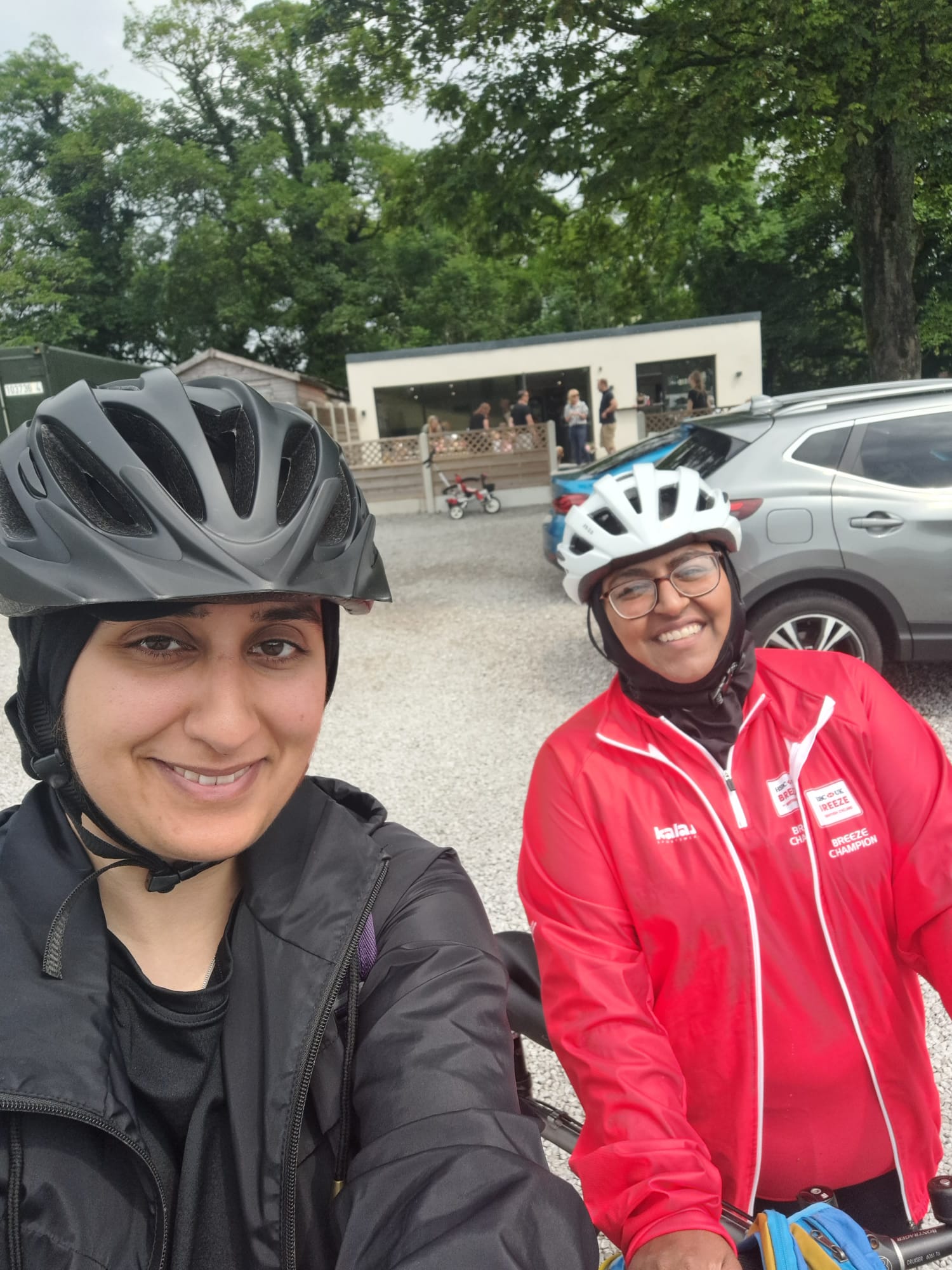 Two women wearing cycling helmets and smiling at the camera. Naida is wearing a red jacket and white helmet, the other lady is wearing all black,
