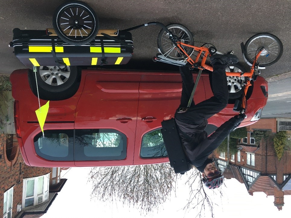 A teenager is sitting on an orange Brompton folding cycle smiling at the camera. They have an instrument case on their back and are towing a flatbed trailer with another very large instrument case strapped to it.