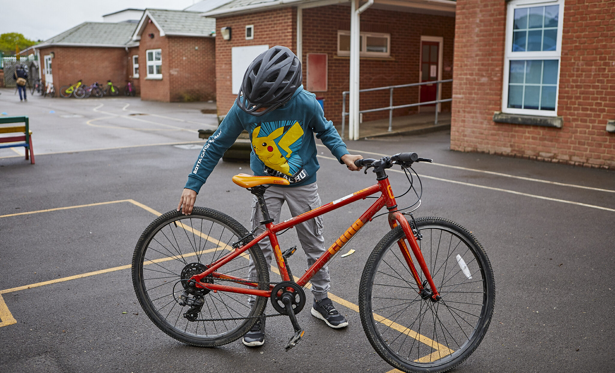 A young boy in a playground checking his bicycle tyres. His bike is red and he is wearing a green and yellow jumper and a bike helmet.