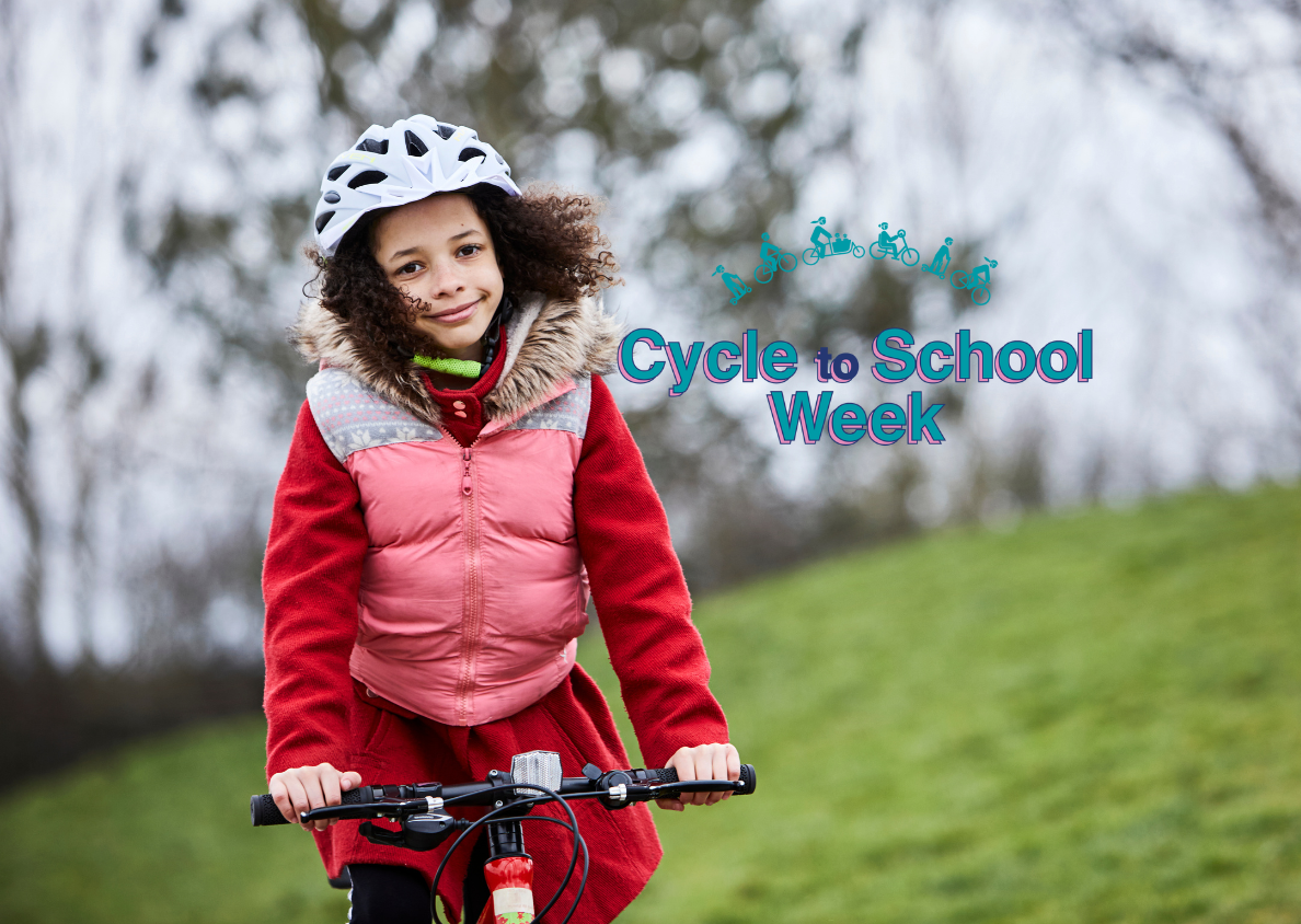 A young schoolgirl is riding her bicycle to school. She is facing head-on to the camera, both hands on the handlebars, and smiling. She is wearing a helmet, and grass can be seen in the background. The logo for Cycle to School Week is visible.