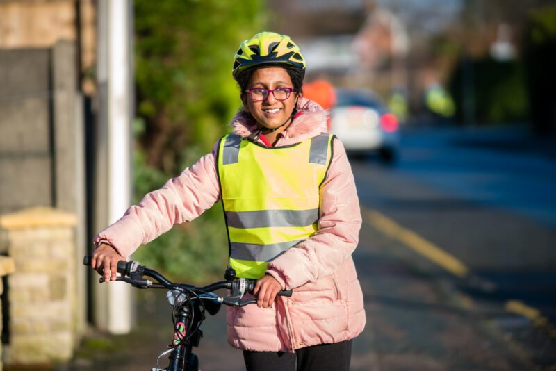 A woman is pushing her cycle. She is wearing a pink coat with a hi-vis tabard over the top.