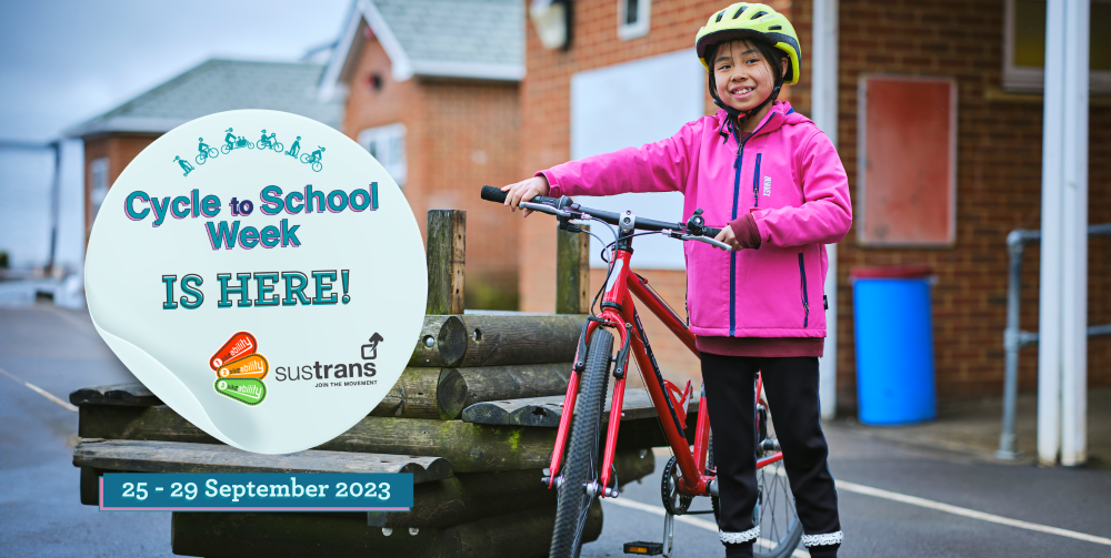 A young girl in a pink jacket is standing with a bicycle in a playground. She has a big grin on her face. There is a sticker on the image that says Cycle to School week is here!
