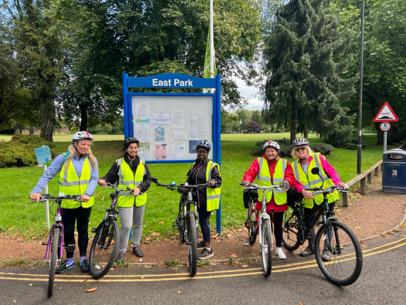 A group of five women with bikes are standing in a park. They are wearing hi vis and smiling