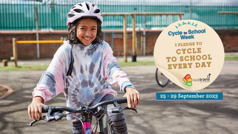 A girl on her cycle in a playground smiling. There is a sticker on top saying I pledge to cycle to school every day