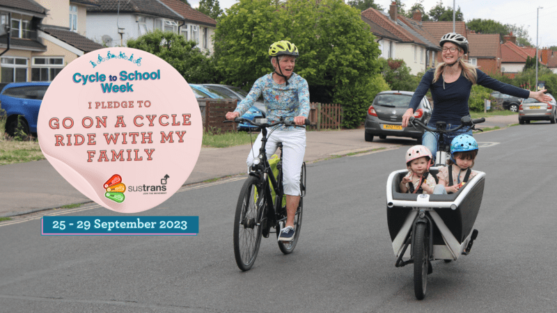 Two women on cycles, one with two little children in a front carrier. There is a sticker on top saying I pledge to go on a cycle ride with my family