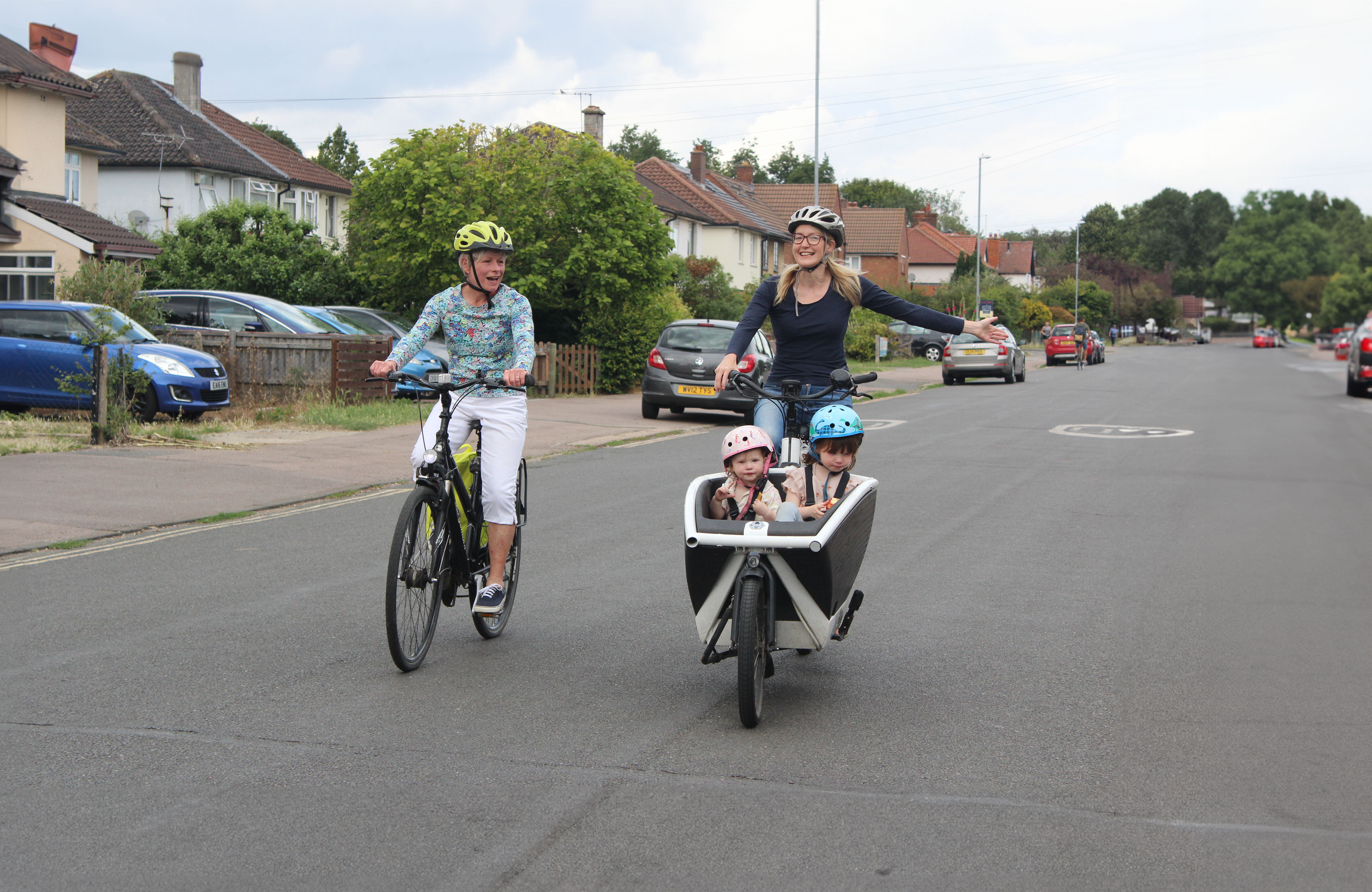Two women are riding their cycles in primary position down a residential road. One is riding a bicycle, while the other is signalling to turn, and riding a cargo cycle, with two young children sitting in the front compartment. They are all wearing safety helmets and smiling.