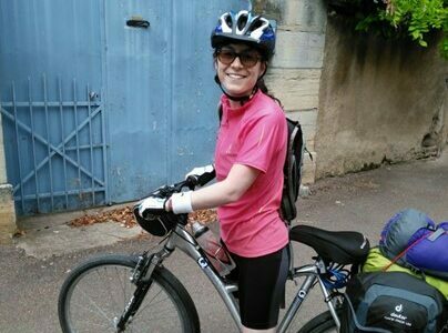 A young woman with sunglasses wearing a pink t-shirt, black shorts and white trainers stood in a road on a grey cycle with bags on the back wearing a blue and silver helmet. In the background there is a blue gate, white wall and a tree.