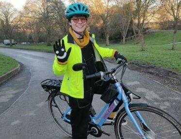 Lucy on her blue e-cycle wearing a hi vis jacket, smiley face gloves, a blue helmet, yellow jumper and socks and black dungarees and trainers with a road and park in the background full of trees