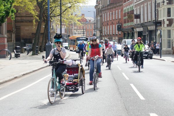 Lucy on her mint green converted e-cycle wearing a white t-shirt, black dungarees and a blue helmet with a black toy cat strapped on the top. Behind her is a group of people of all ages cycling together as a big group on a city centre road with buildings and trees on either side.