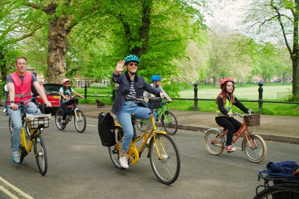 Lucy on her now yellow converted e-cycle wearing a striped blue and white shirt, blue jacket and jeans with a blue helmet riding on the road. She is waving to the camera. Next to her are other people cycling and there is a park with trees and grass in the background.