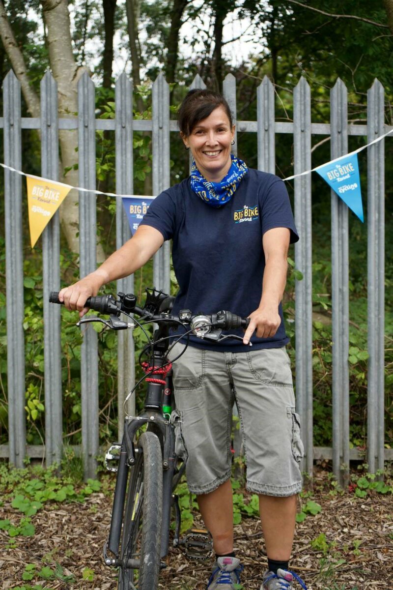 Michelle is standing in front of a fence covered in bunting with her bicycle. She is wearing a dark blue t shirt and grey shorts and smiling