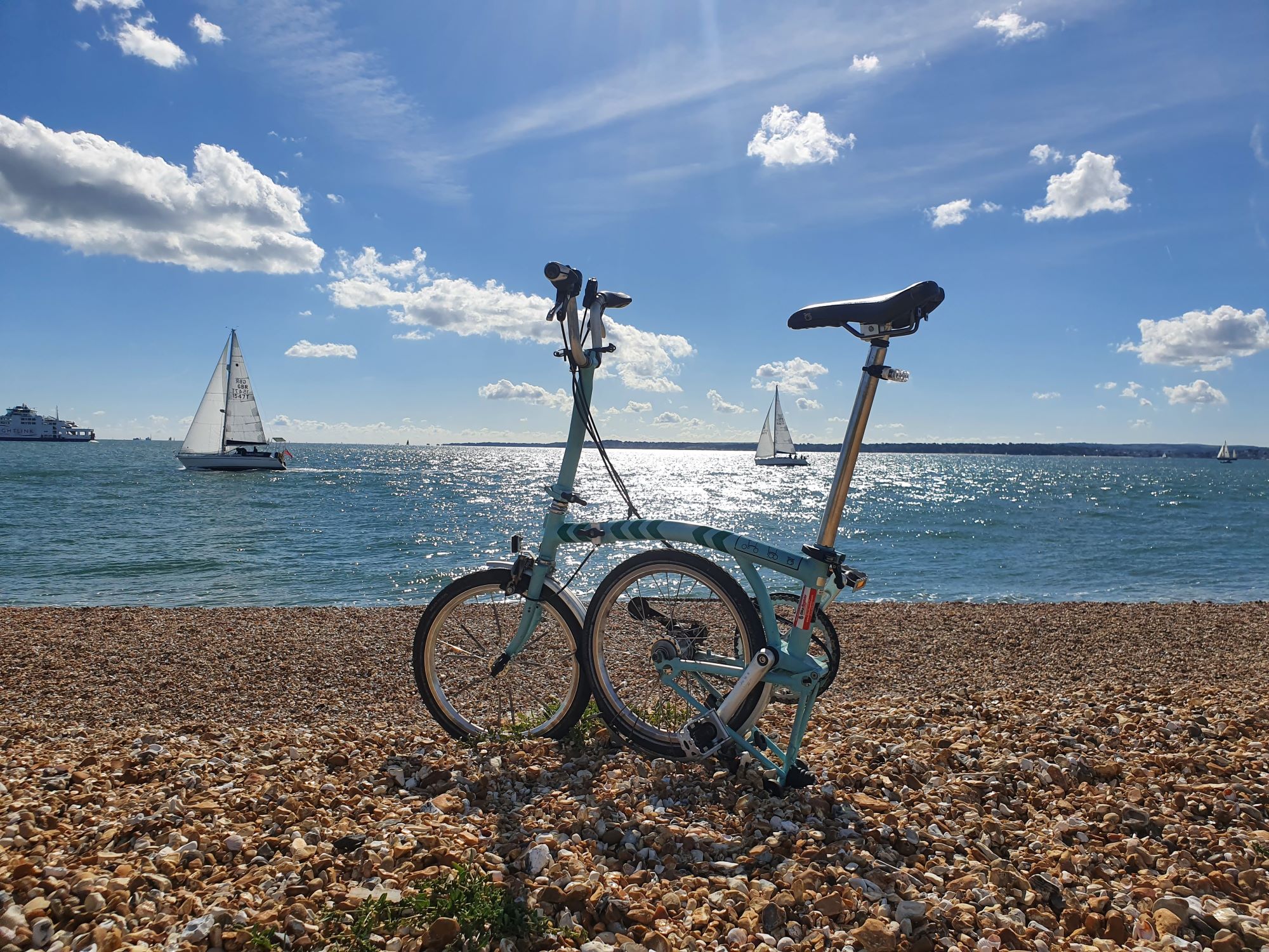 A folding bicycle on the beach with a sail boat on the sea in the background