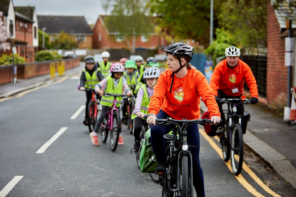 Two Bikeability instructors are leading a group of students as they cycle on a road. They are waiting at a junction to turn