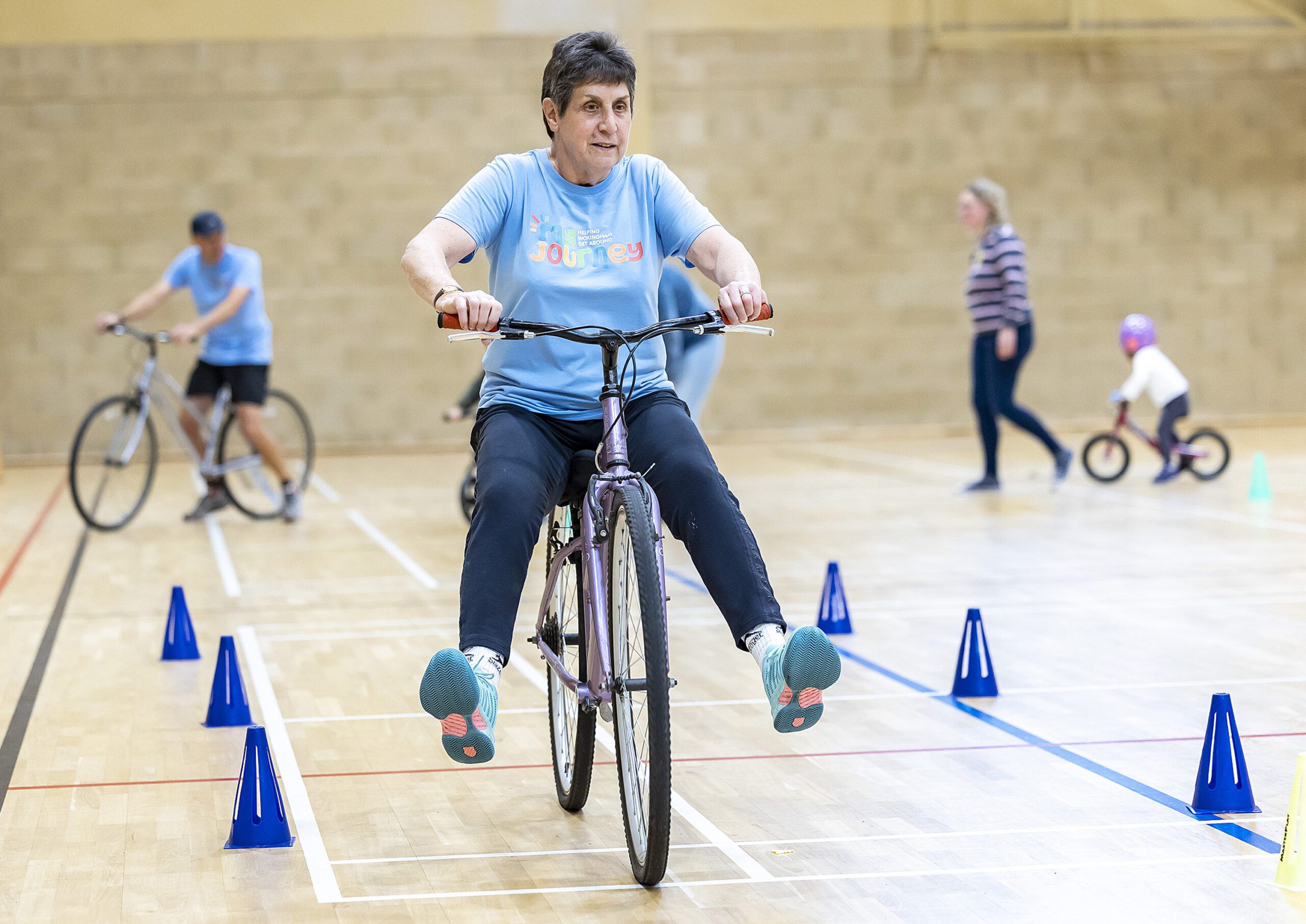 A woman on a bicycle with her legs up as she enjoys gliding through some cones in a gym