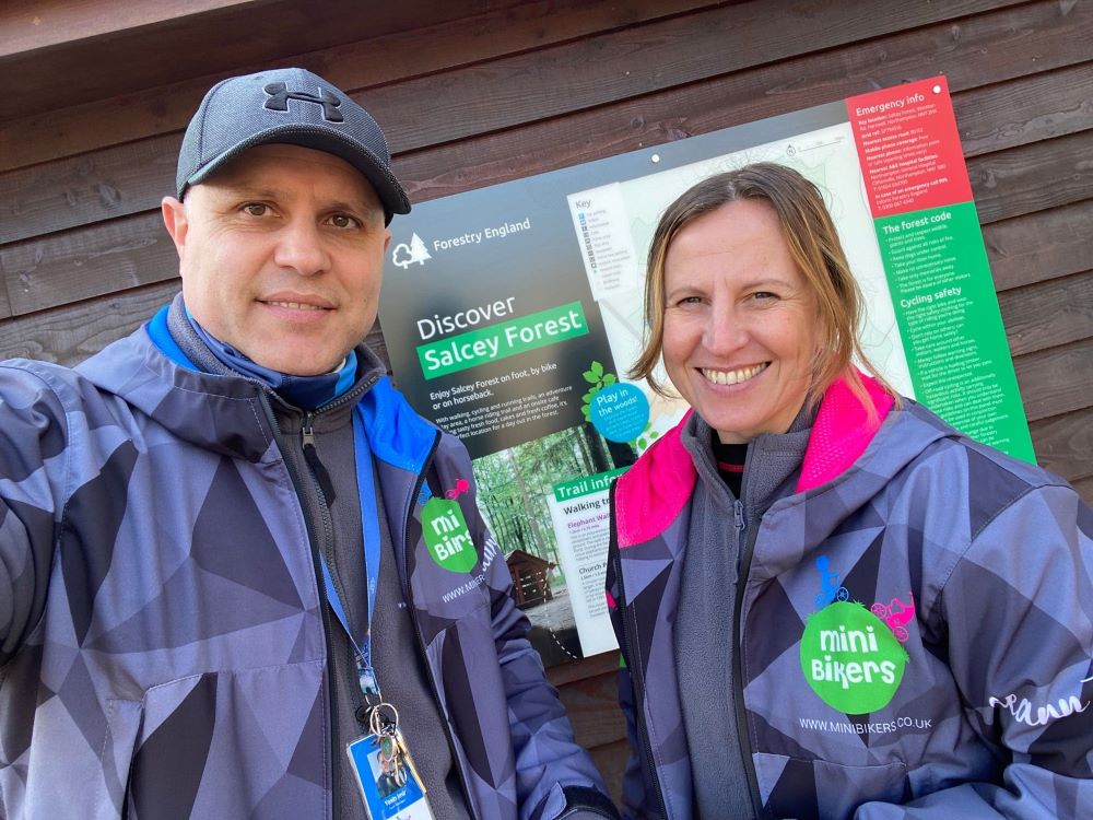 Two people are smiling at the camera. They both have blue jackets with a logo that says Minibikers