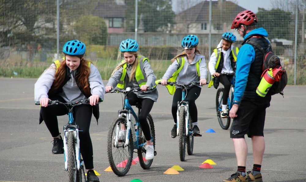A group of school children cycling around cones in a playground while a Bikeability instructor gives tips