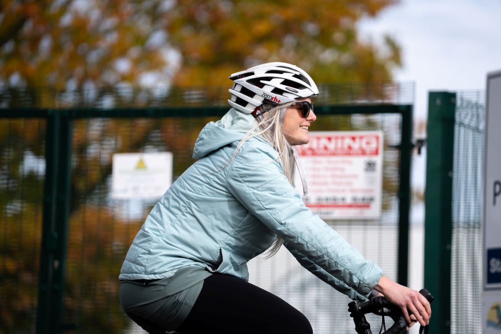 A woman wearing a light blue coat and a white and black cycling helmet cycling past a green gate