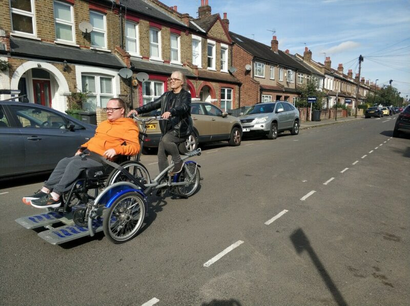 Two people using a cycle with a space for a wheelchair to ride in the front