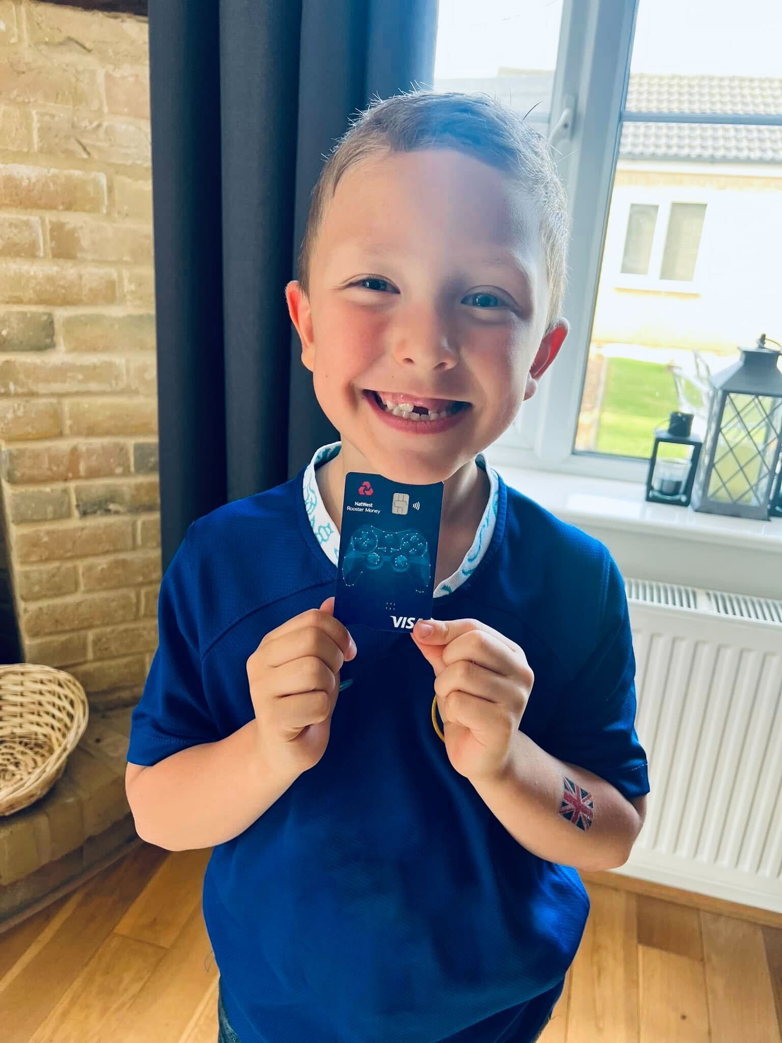 A young boy in a football top is smiling and holding up a rooster money card