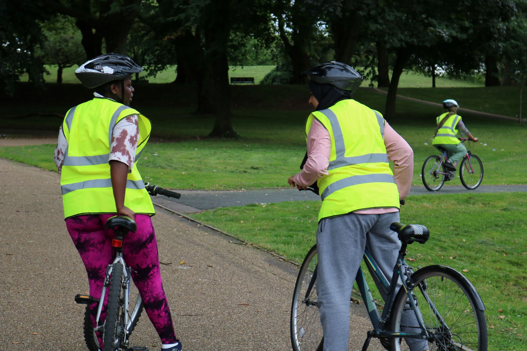 Two women in hi-vis jackets on cycles in a park. They are facing away from the camera