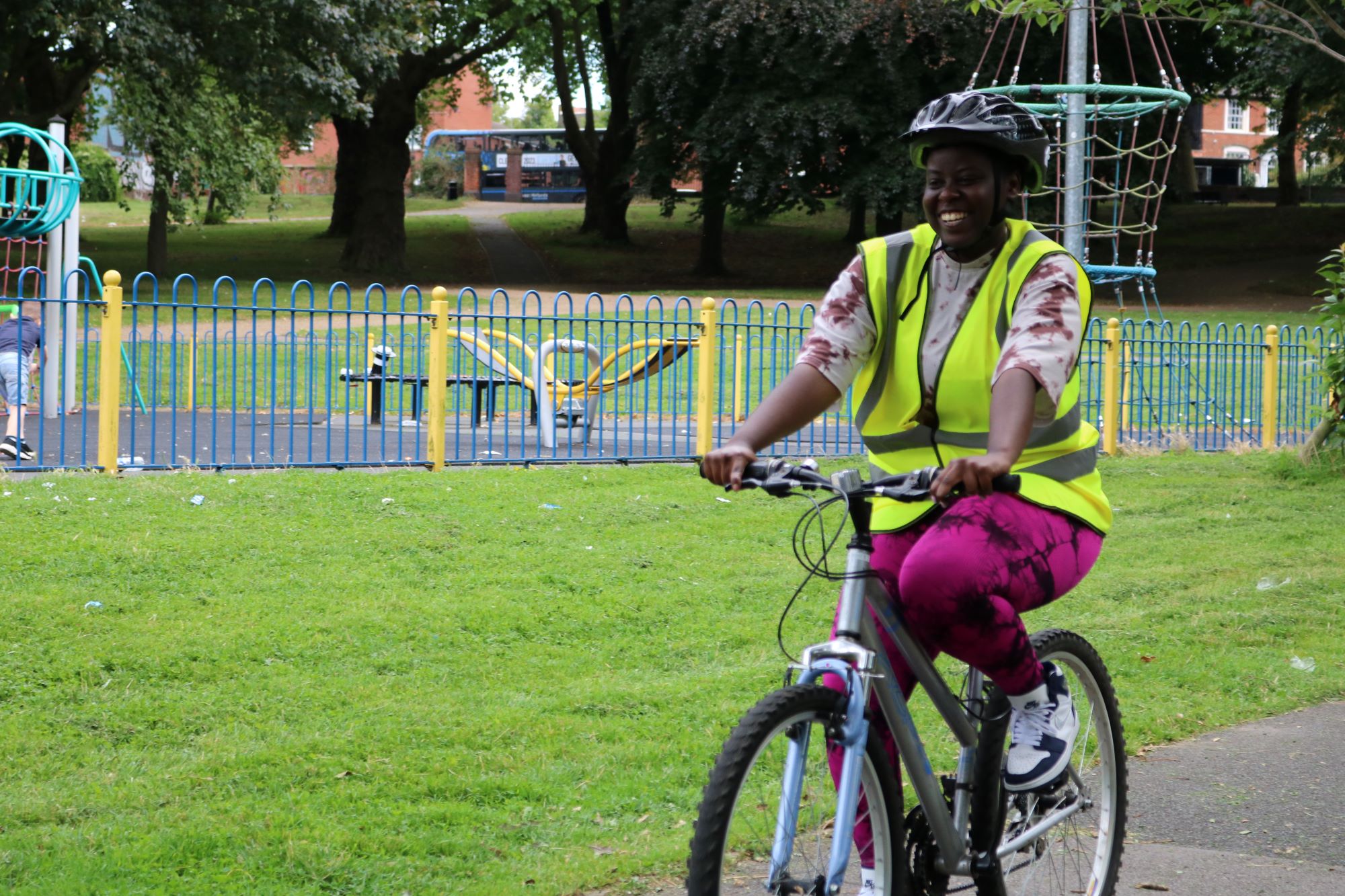 A woman cycling in a park in front of a playground. She is smiling