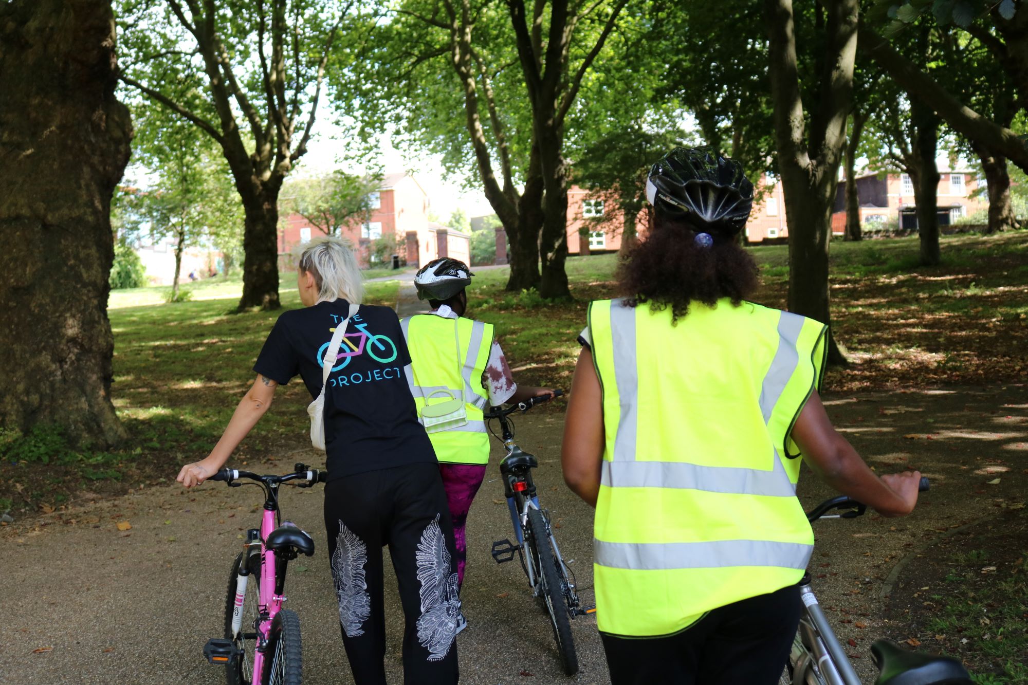 Three women pushing their cycles in a wooded area. They are wearing hi-vis vests and walking away from the camera