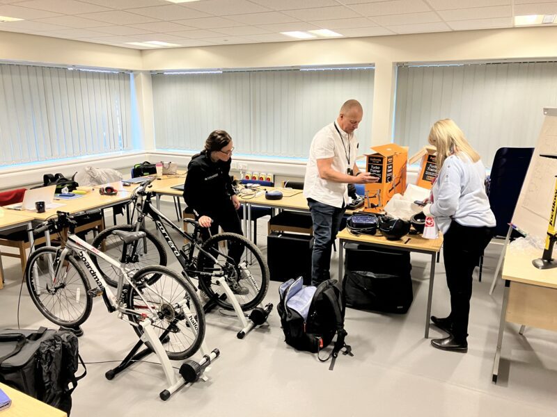 Three people in a room are setting up equipment including computers and bicycles on stationary turbo trainers