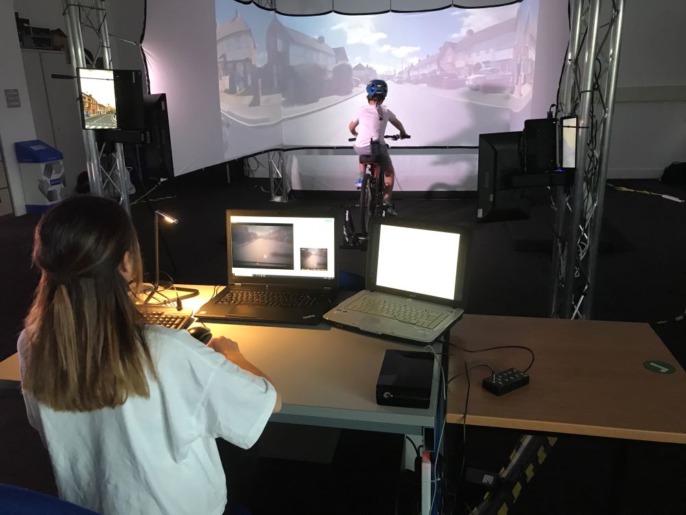 A child is wearing a headset and riding a stationary bike indoors. He is surrounded by screens showing an on road route. A woman is behind a desk of laptops watching on.