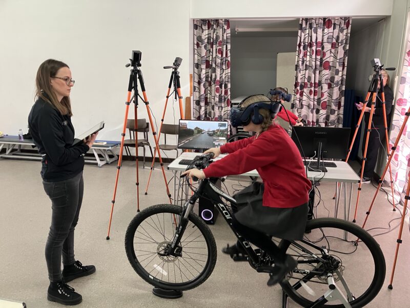 A child in a school uniform is on a stationary cycle in a a lab while a woman stand in front of them with a clipboard