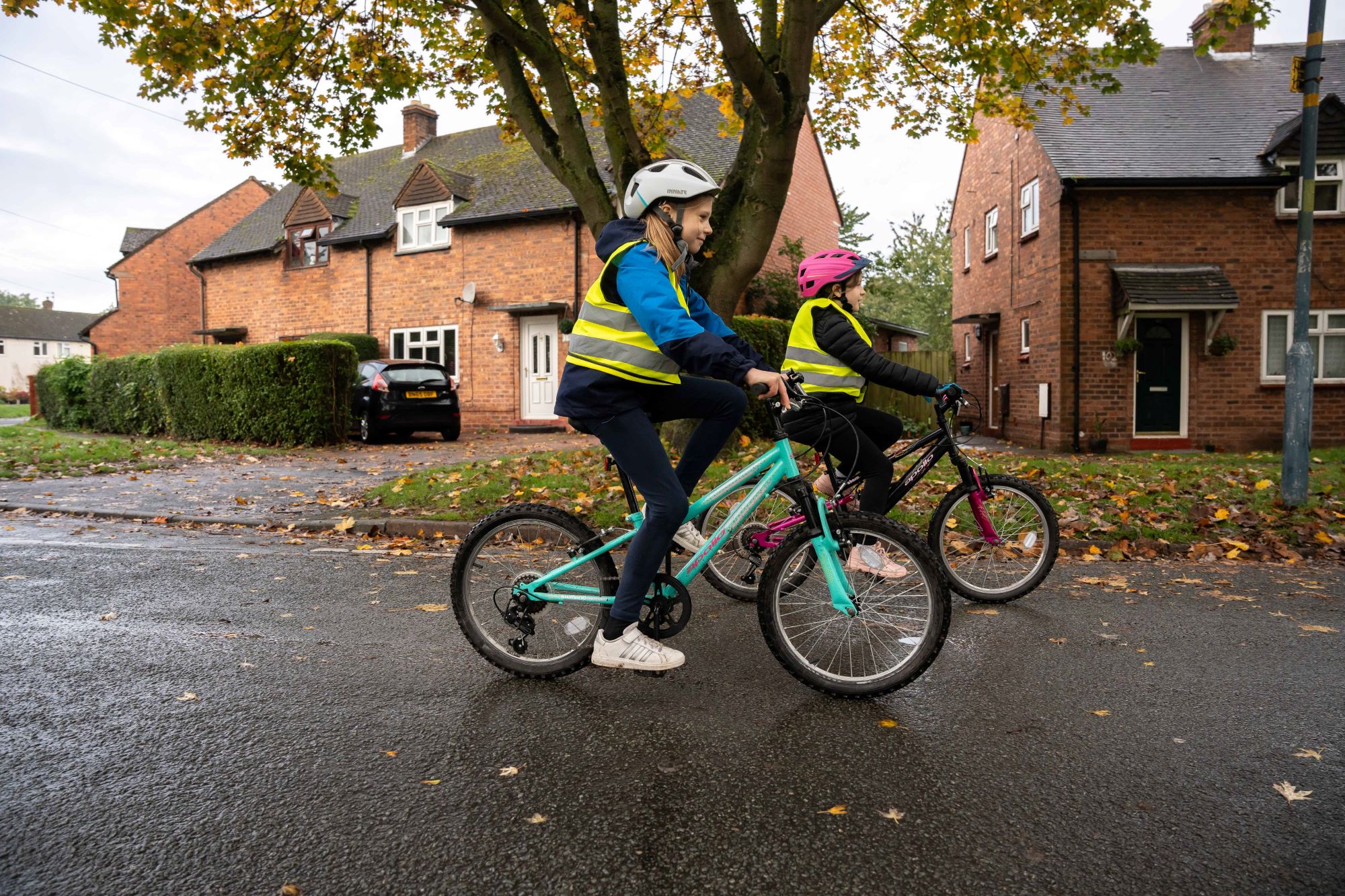 Two girls cycling on a road. They are wearing hi vis vests and helmets