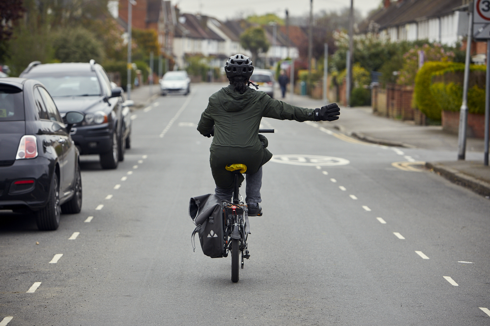 An adult is cycling is cycling to work down a residential road. She is wearing a safety helmet, has a bag attached to her cycle, and is signalling to turn right.