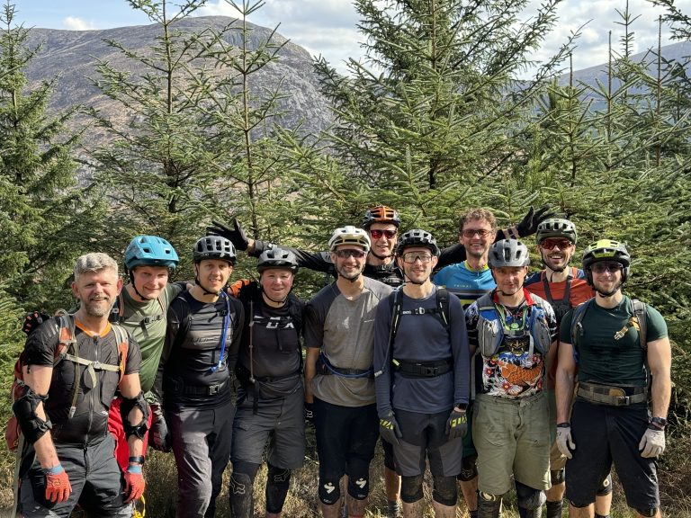 A group of Out MTC members stand in a forest. They are all wearing cycling helmets and riding gear.